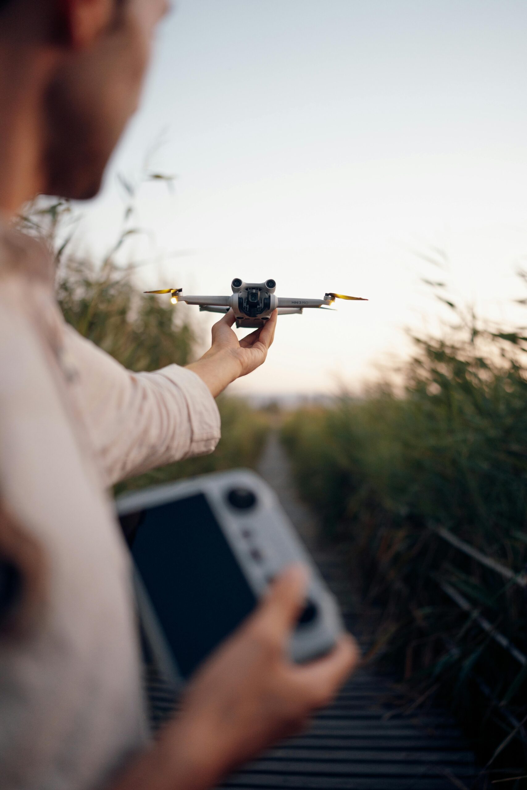A man flying a drone using a handheld controller in a natural outdoor setting at dusk.
